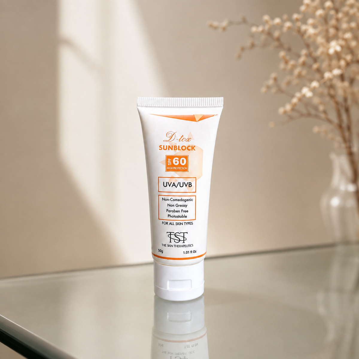 A white tube of TSI Sunblock SPF 60 UVA/UVB sunscreen stands on a reflective glass surface. The background is a warm beige wall with soft sunlight shadows and a blurred vase of dried flowers on the right.